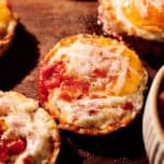 Three baked Breakfast Pizza Egg Bites topped with melted cheese and tomato sauce are displayed on a wooden surface. The muffins have golden edges and a slightly crispy appearance.