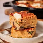 Two square slices of baked pasta frittata casserole with visible herbs and cheese are stacked on a plate with a fork. A larger baking dish with more pasta frittata is in the background.