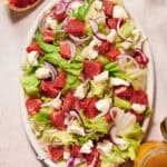 Oval plate of summer salad with iceberg lettuce, grapefruit segments, red onion slices, and chunks of cheese on a light surface. Grapefruit halves and small jars are visible nearby.