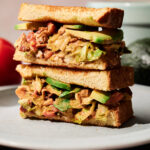 A close-up of two sandwich halves stacked on a plate, filled with lettuce, avocado slices, and a savory filling between toasted bread slices. An avocado and tomato are in the background.