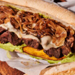 A Beef Stew Sandwich on a sub roll filled with grilled beef, melted cheese, sautéed onions, green sauce, and a slice of plantain, resting on parchment paper with part of another sandwich visible.