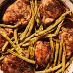 A white pot filled with browned One Pot Chicken Thighs and green beans, cooked in a savory sauce, sits on a wooden surface.