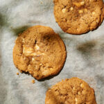 Three large cookies with white chocolate chunks sit on a sheet of parchment paper; one cookie has a bite taken out of it.