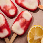 Five red and white popsicles are arranged on a tan surface next to a sliced lemon, with visible wood sticks and bright natural lighting.