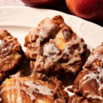 Close-up of apple fritters with white glaze on a plate, with two whole apples in the background.