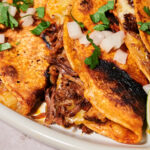 Close-up of crispy birria tacos stuffed with tender shredded beef, topped with chopped onions and cilantro, served on a white plate with lime wedges.