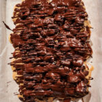 A sheet of parchment paper topped with a layer of nuts and caramel, heavily drizzled with melted dark chocolate and studded with dried cherries and sweet dates, creating a decadent Chocolate Cherry Date Bark dessert.
