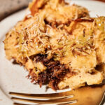 Slice of Dubai chocolate pistachio French toast bake on a plate, showing layers of fluffy challah bread filled with chocolate, drizzled with pistachio cream, and sprinkled with crispy kataifi or filo pastry, served with a gold fork.