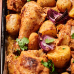 Roasted chicken drumsticks and thighs with golden potatoes, red onion wedges, and fresh cilantro on a baking tray, all seasoned and baked to a crispy, golden finish—perfect for anyone craving delicious roasted chicken packed with flavor.