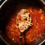 Overhead view of shredded chicken in a tomato-based broth inside an Instant Pot, with a ladle lifting some of the chicken and vegetables from the soup.