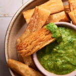 A bowl of crispy Fried Yuca Fries surrounds a small dish of green dipping sauce, with one fry dipped in the sauce.