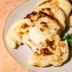 A plate with sliced, golden-brown arepas. One arepa is opened, showing melted cheese stretching out. Some fresh greens are on the side.