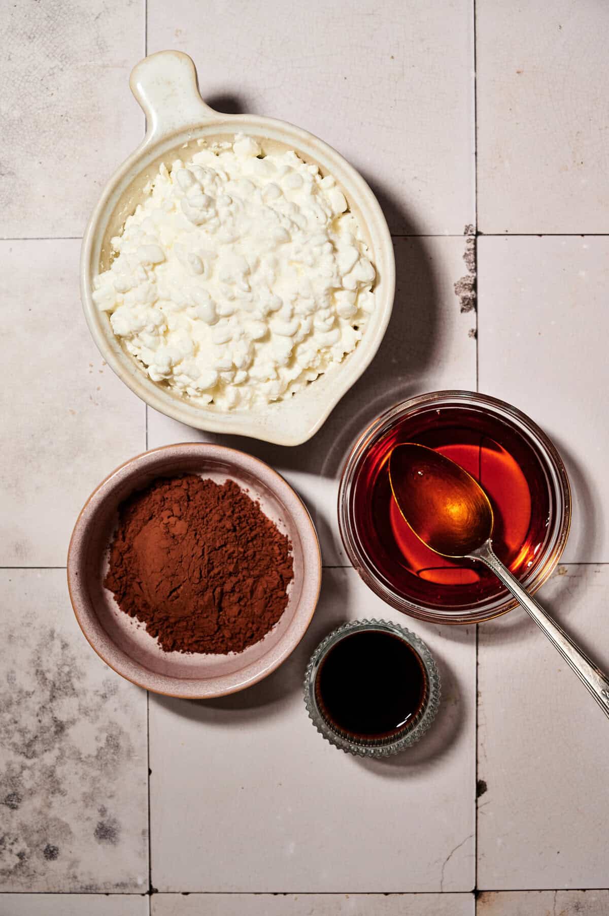 High protein chocolate mousse preparation with cottage cheese, unsweetened cocoa powder, maple syrup, and vanilla extract, arranged on a tiled surface.