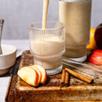 A creamy smoothie is being poured into a ribbed glass. Another full glass sits beside it. Apple slices and cinnamon sticks are on the tray nearby, with apples in the background.