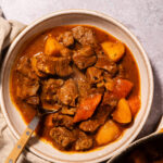 A bowl of beef stew with chunks of beef, potatoes, and carrots in a rich brown broth, with a spoon resting inside the bowl. A beige napkin is beside the bowl on a gray surface.