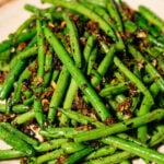 A plate of sautéed green beans garnished with crispy bits of garlic and herbs, served on a light-colored dish atop a wooden surface.