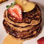 A stack of Protein Oat Pancakes topped with a sliced strawberry and a piece of golden kiwi, drizzled with syrup, on a white plate.