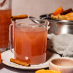 A glass mug of pinkish cider garnished with a cinnamon stick sits on a plate beside tangerines, a pitcher of cider, a bowl with fruit, and a gray cloth.