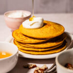 A stack of pancakes topped with whipped cream, with honey being drizzled on top. Surrounding the plate are a bowl of honey, chopped nuts, a fork, and a small bowl in a bright kitchen setting.