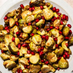 A plate of roasted Brussels sprouts topped with fresh pomegranate seeds, with a small bowl of red pepper flakes in the background.