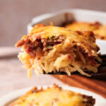 A close-up of a cheesy casserole slice with ground meat and vegetables being served, showing melted cheese and shredded potatoes. The rest of the casserole is visible in the background.