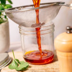(oil being strained into jar): Straining homemade achiote oil through a fine mesh sieve into a glass jar, capturing its vibrant red/orange hue.