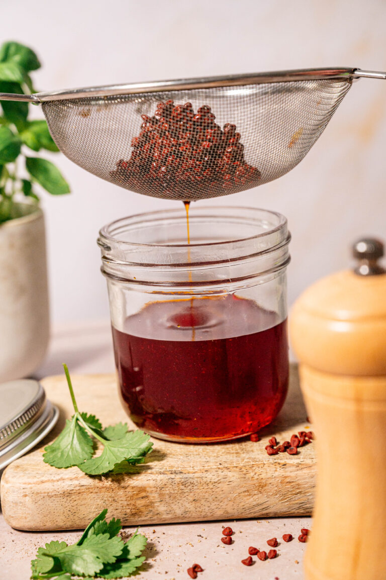 Annatto seeds resting in a strainer after infusing achiote oil, with finished oil collected in a glass jar below.