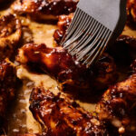 A silicone brush spreads barbecue sauce on baked chicken wings, which are resting on a parchment-lined baking sheet, highlighting the irresistible appeal of perfectly baked chicken wings.