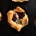 Close-up of mini puff pastry tarts filled with goat cheese and dark fruit preserves, garnished with a sprig of rosemary, baked in a black muffin tin.