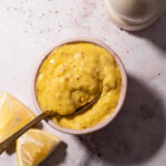 Close-up overhead shot of a spoon dipping into a bowl of creamy homemade mayonnaise, highlighting rich yellow color, grainy mustard flecks, and fresh lemon wedges on the side.