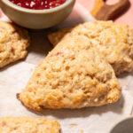 Golden, flaky cottage cheese scones on parchment paper with a bowl of red jam nearby, set on a wooden board and pink surface.