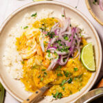 A bowl of white rice topped with yellow curry shrimp reminiscent of Sango de Camaron, sliced red onions, chopped herbs, and a lime wedge, with a fork resting inside the bowl. Lime halves and bowls are visible nearby.