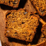 Close-up of sliced banana bread with visible walnuts, placed on a wooden cutting board. The bread looks moist and textured, with a golden-brown crust and scattered nut pieces throughout.