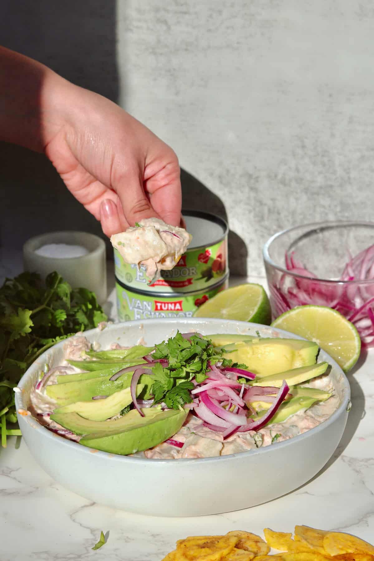 Holding up a crispy plantain chip above a bowl of Ecuadorian ensalada de yuca y atún, showing the creamy tuna and yuca salad with cilantro, lime, and ingredients displayed in the background.