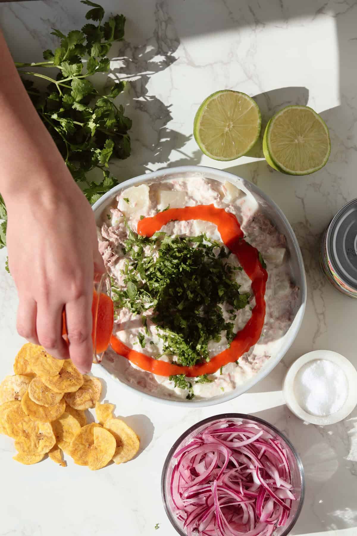 Step-by-step photo of ensalada de yuca y atún showing a bowl of creamy yuca tuna salad being topped with chopped cilantro and a swirl of tomate de árbol hot sauce, surrounded by limes, red onion, and plantain chips.