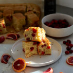 Stack of blood orange cranberry olive oil cake squares on a white plate with fresh blood orange slices around the dessert.