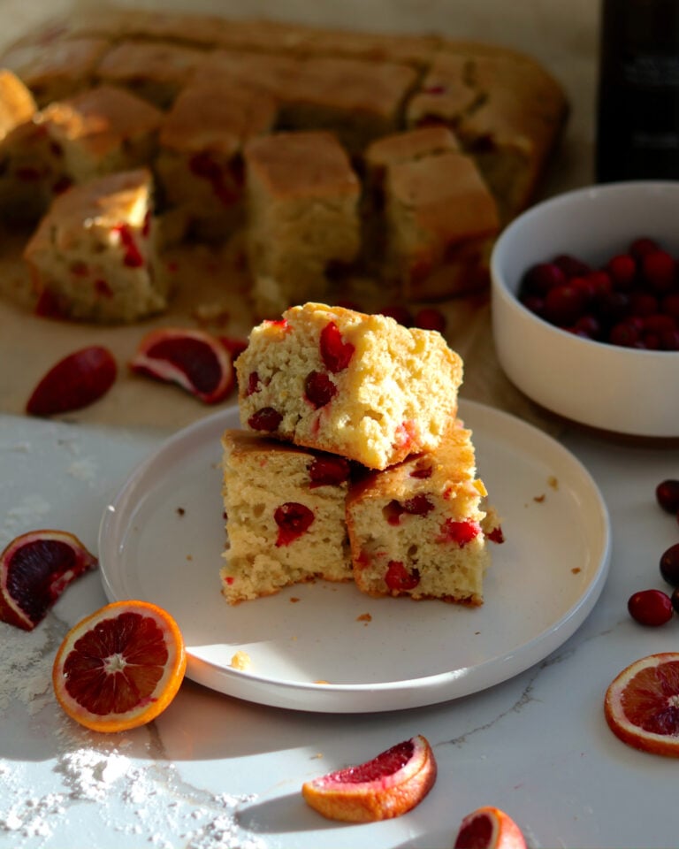 Blood orange olive oil sheet cake cubes on a plate, surrounded by scattered cranberries and sliced blood oranges on a marble surface.