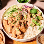 A bowl of rice topped with cooked chicken, sliced cucumber, red onion, avocado, fresh cilantro, sesame seeds, and a creamy sauce, with extra cilantro and sauce in small bowls nearby.