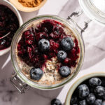 A jar of overnight oats topped with blueberries and berry compote, surrounded by bowls of blueberries and a small bowl of cinnamon on a marble surface.