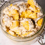 A glass jar filled with overnight oats, topped with chunks of fresh mango and a sprinkle of shredded coconut, with a spoon inside.