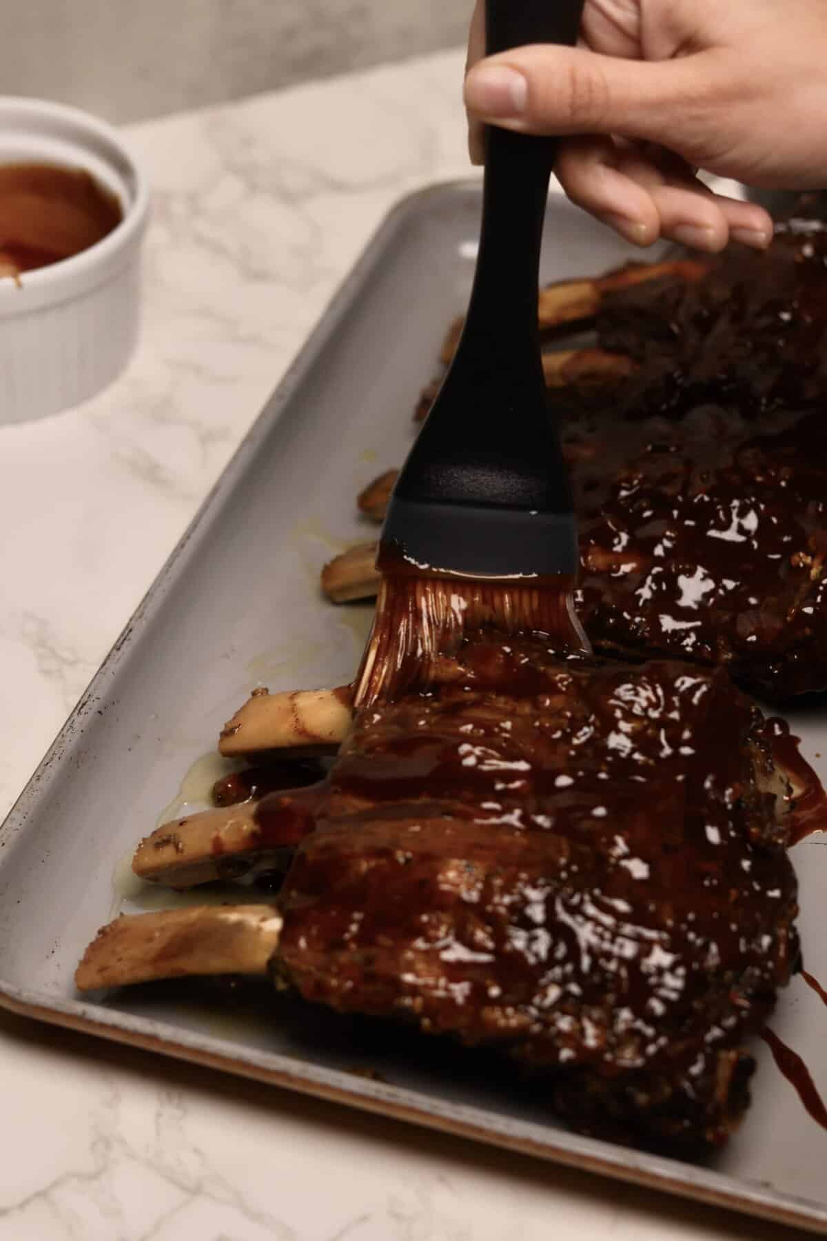 Close-up of cooked beef back ribs on a sheet pan being brushed with BBQ sauce to coat evenly before going under the broiler.