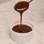 A spoon drizzles thick Organic Barbecue Sauce into a small white ramekin, which is filled with the same glossy sauce, on a light marble surface.