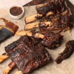 Close-up of broiled crockpot baby back ribs on a parchment-lined board, with several ribs sliced apart to show tender meat, a basting brush, and a small ramekin of extra BBQ sauce.