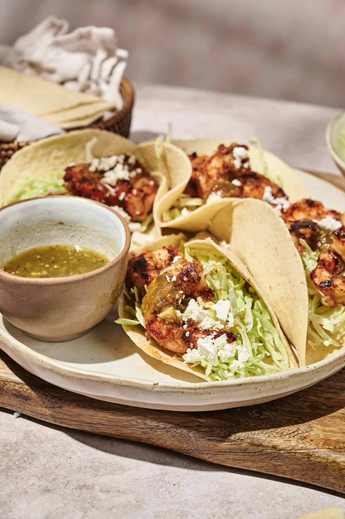 Seared spice-rubbed shrimp tacos in warm corn tortillas filled with shredded cabbage, crumbled cheese, and served with a bowl of salsa verde on a ceramic platter.