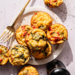A plate of mini egg muffins with spinach, red peppers, and cheese sits beside a fork and pepper grinder on a white surface.