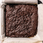 A square pan lined with parchment paper holds a batch of freshly baked chocolate brownies with a rich, slightly textured surface.