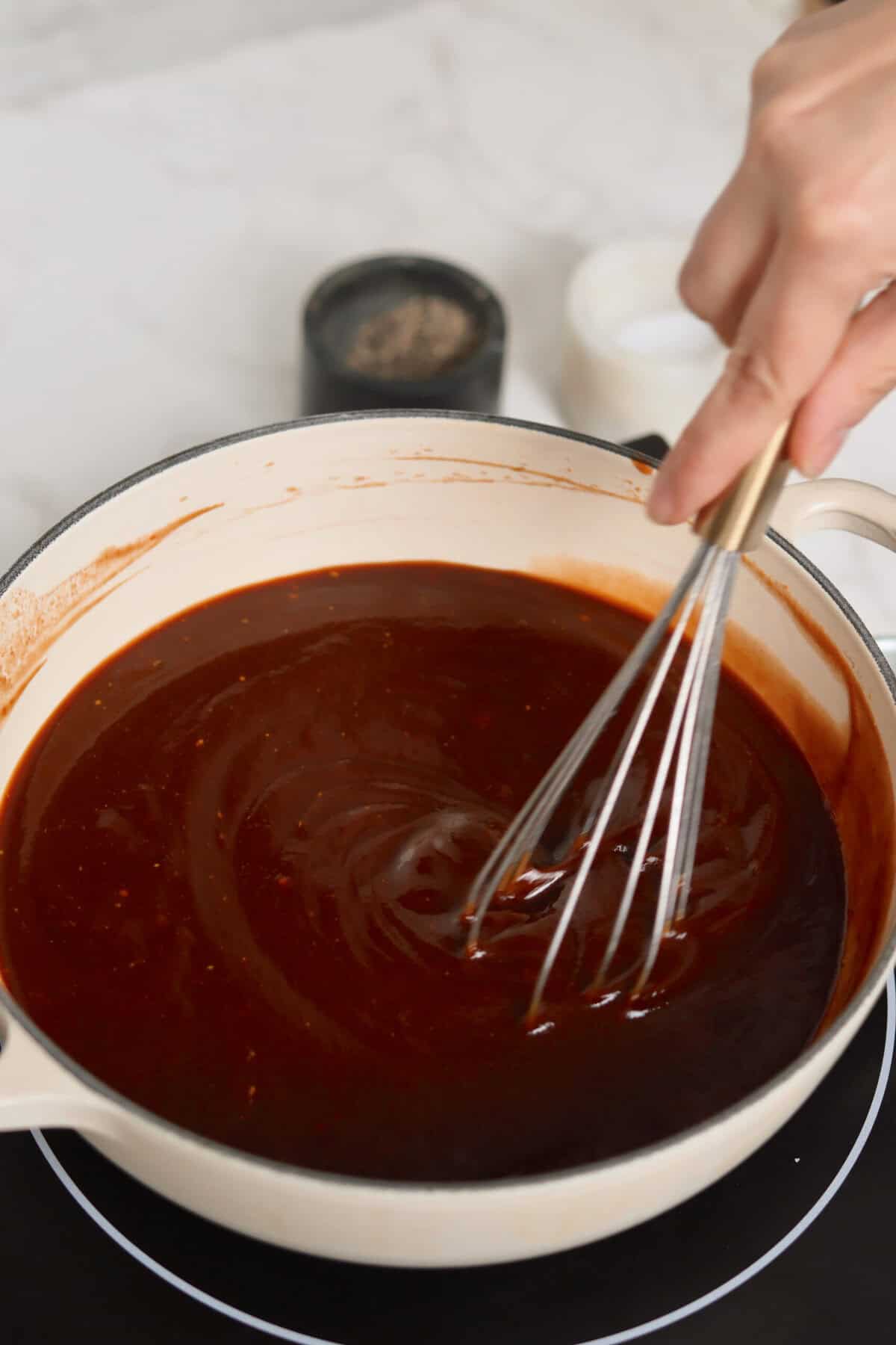 Whisking thick homemade apple cider barbecue sauce in an enameled pot on the stovetop.