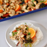 A plate of white rice topped with baked miso chicken, broccoli, and orange slices. In the background is a baking tray filled with more miso chicken, vegetables, and orange slices—a vibrant Miso Chicken sheet pan dinner.
