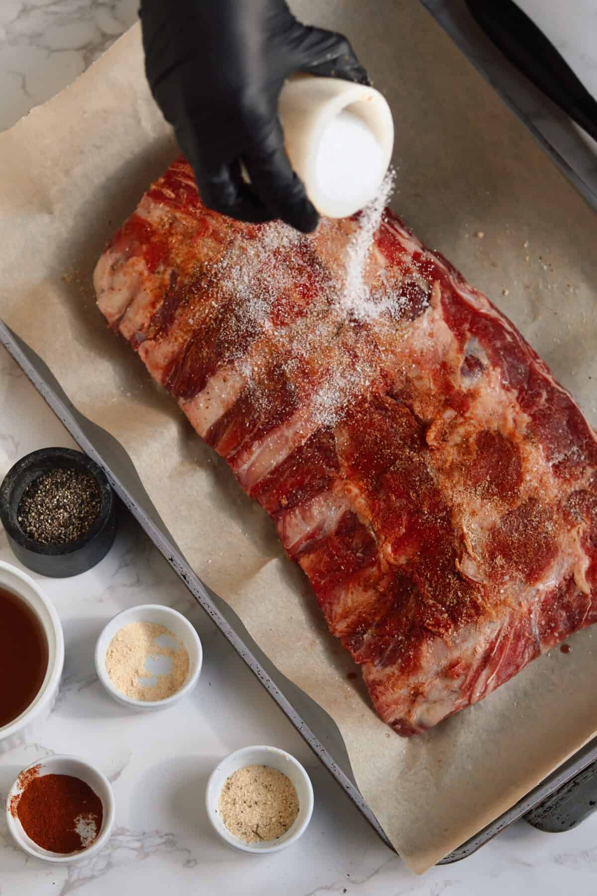 Overhead shot of raw baby back ribs on a parchment-lined sheet pan being sprinkled with salt, surrounded by small bowls of beef broth, paprika, garlic powder, onion powder, and black pepper.
