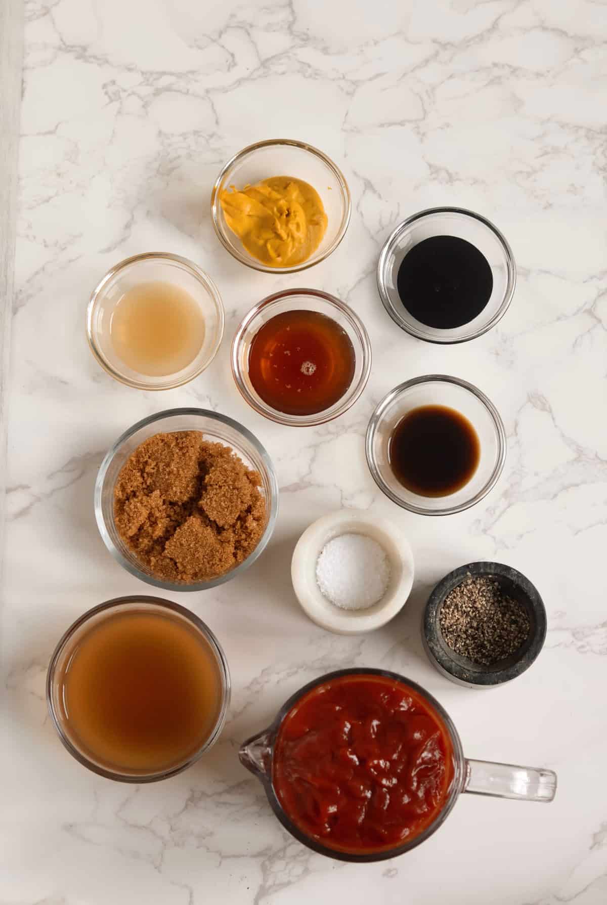 Overhead shot of measured ingredients for apple cider barbecue sauce, including ketchup, vinegar, brown sugar, mustard, honey, Worcestershire, salt, and pepper in glass bowls on a marble surface.